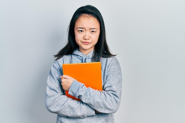 Young chinese girl holding book skeptic and nervous frowning upset because of problem negative person
