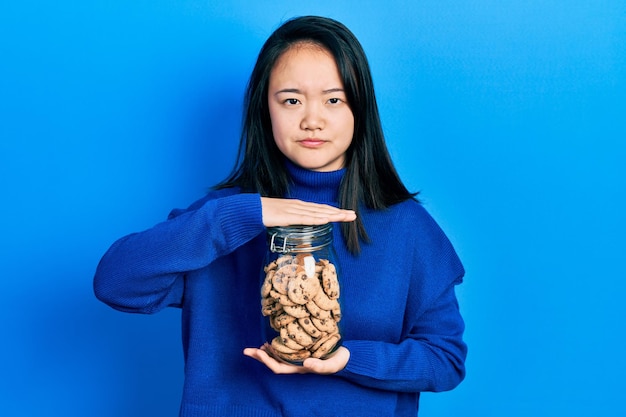 초콜렛 칩 쿠키 병을 들고 있는 젊은 중국 소녀 회의론자와 신경질적인 찡그린 얼굴 때문에 부정적인 문제 때문에 화가 났습니다
