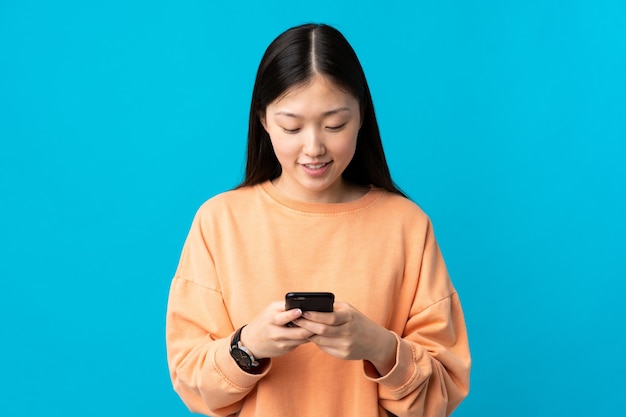 Young Chinese girl over isolated blue sending a message with the mobile