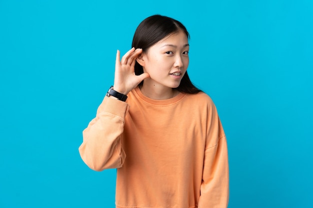 Young Chinese girl over isolated blue wall listening to something by putting hand on the ear