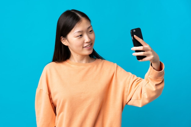 Young Chinese girl over isolated blue wall making a selfie