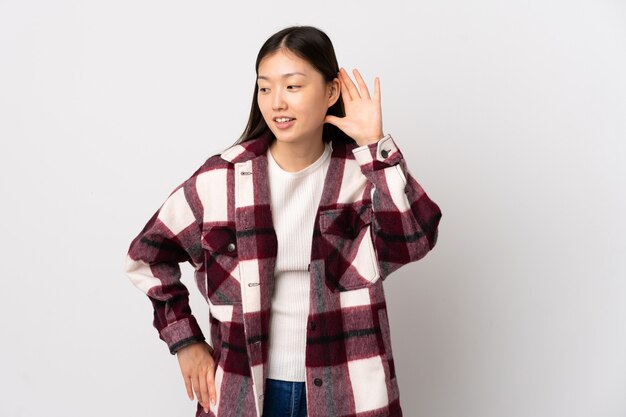 Young Chinese girl isolated listening to something by putting hand on the ear