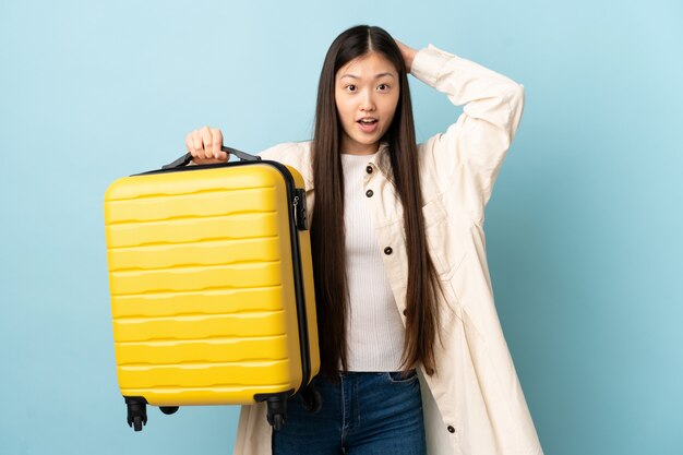 Young Chinese girl over isolated wall in vacation with travel suitcase and surprised