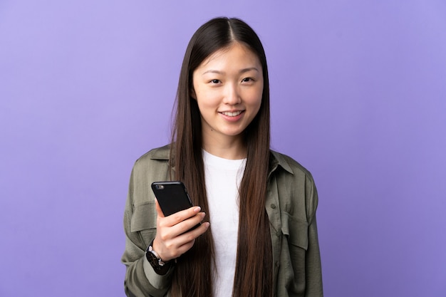 Young Chinese girl using mobile phone over isolated purple smiling a lot