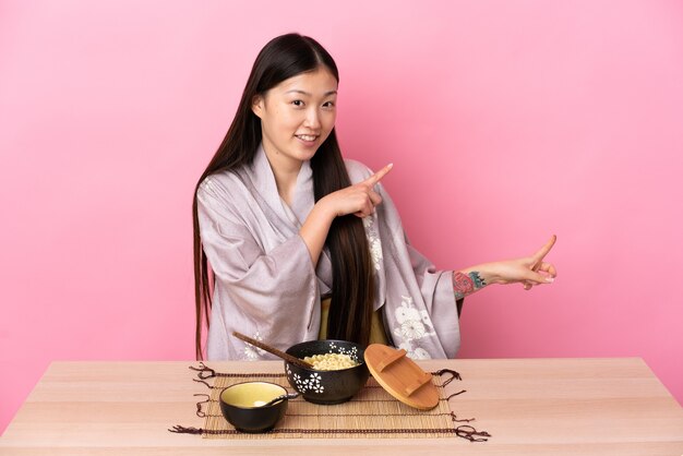 Giovane ragazza cinese che indossa il kimono e mangia le tagliatelle che punta il dito di lato e presenta un prodotto