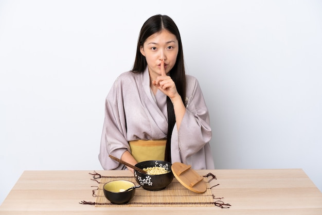 Молодая китаянка в кимоно и ест лапшу, показывая знак тишины, кладя палец в рот