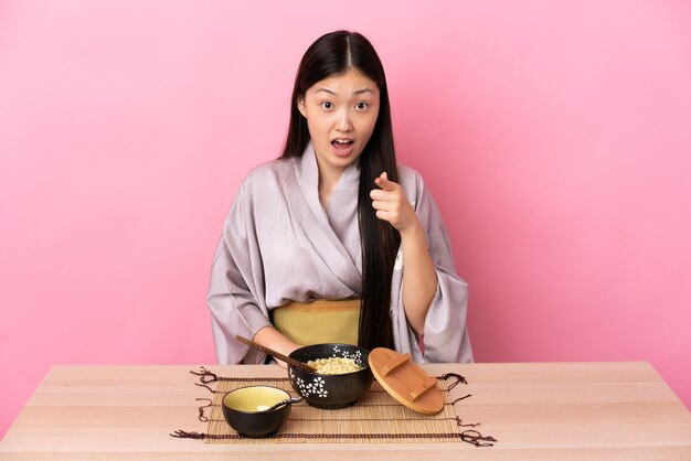 Giovane ragazza cinese che indossa il kimono e mangia le tagliatelle sorpreso e indicando la parte anteriore