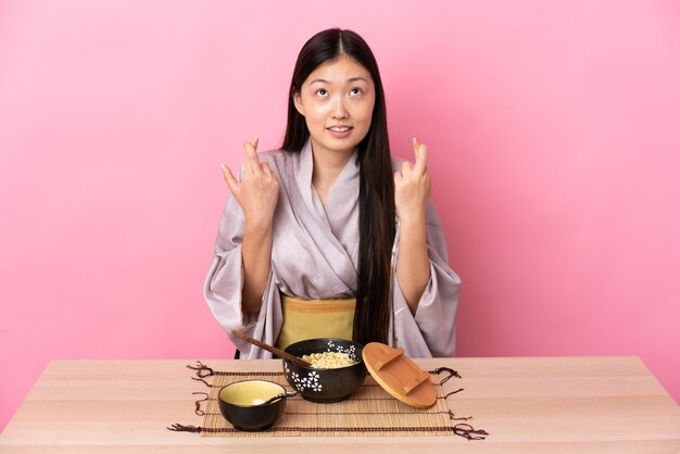 Young Chinese girl wearing kimono and eating noodles with fingers crossing and wishing the best