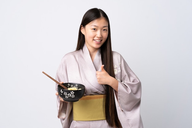 Young Chinese girl wearing kimono over isolated white wall with thumbs up because something good has happened while holding a bowl of noodles with chopsticks