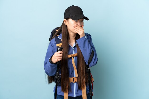 Young Chinese girl with backpack and trekking poles over isolated blue background covering mouth and looking to the side