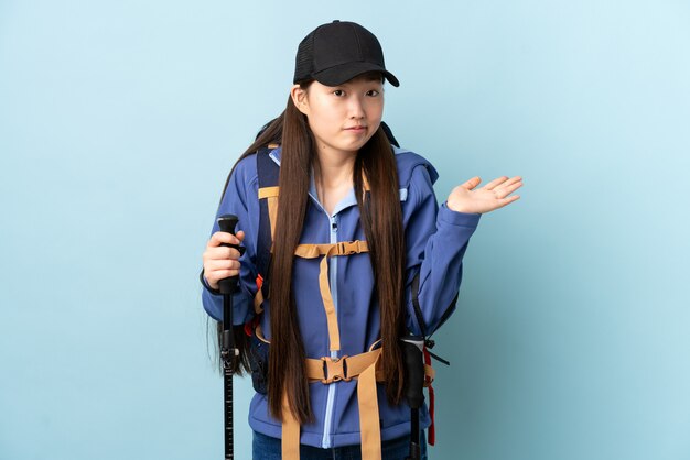 Young Chinese girl with backpack and trekking poles over isolated blue having doubts while raising hands