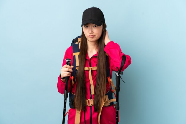 Young Chinese girl with backpack and trekking poles over isolated blue having doubts