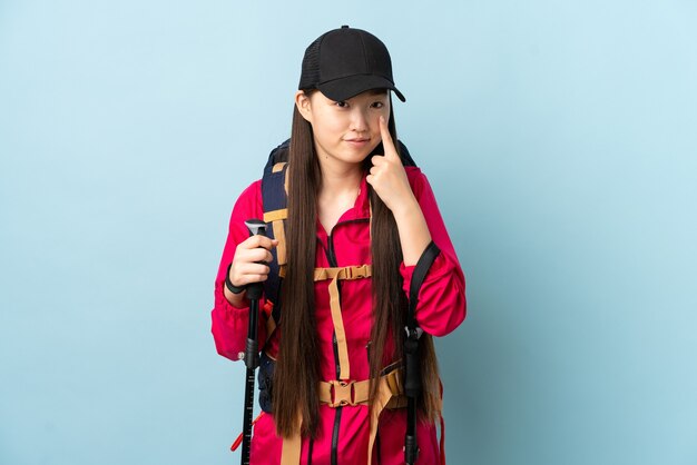 何かを示す孤立した青の上にバックパックとトレッキングポールを持つ若い中国の女の子