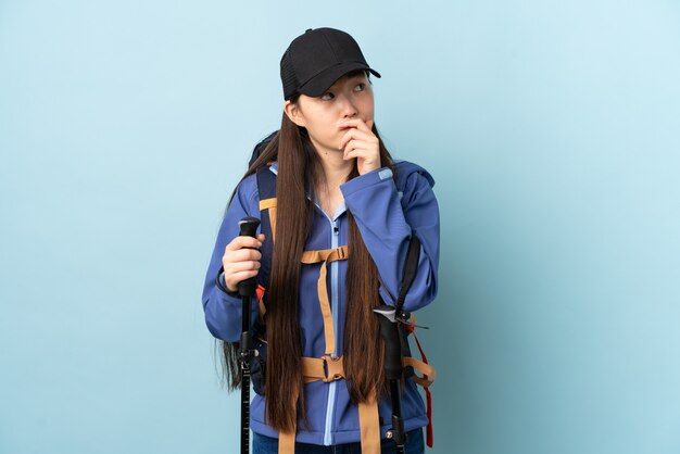 Молодая китайская девушка с рюкзаком и треккинговыми палками над синей стеной, у которой есть сомнения и смутное выражение лица