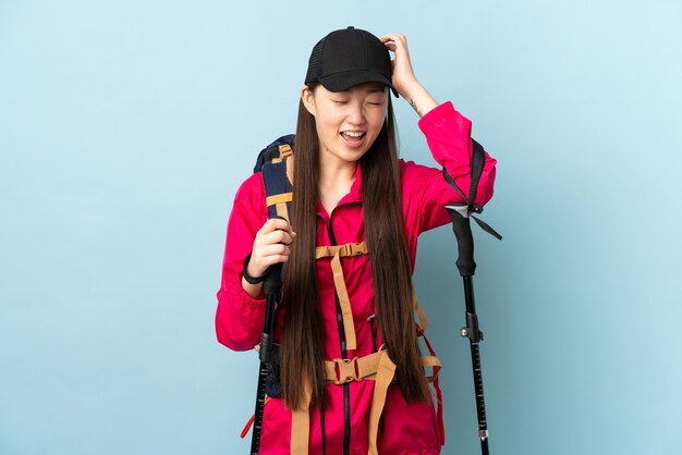 Young Chinese girl with backpack and trekking poles over isolated blue wall laughing