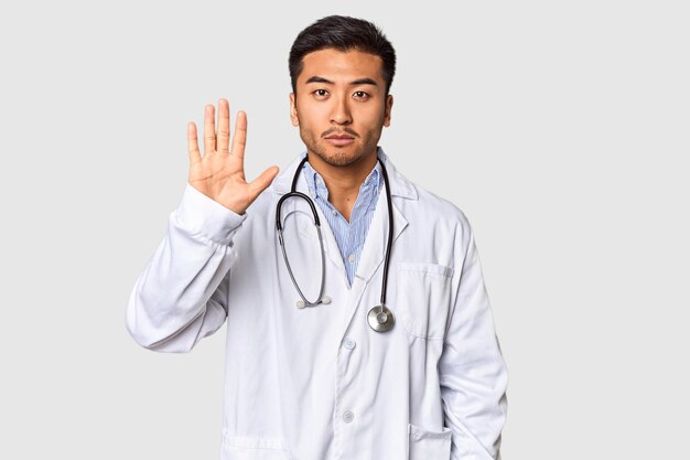 젊은 중국인 의사가 스튜디오에서 미소 짓고 기 ⁇ 게 손가락으로 5번을 보여줍니다.