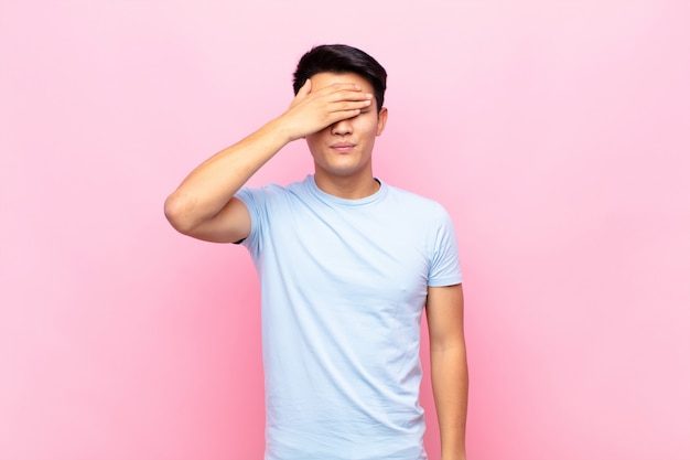 Young chinese man covering eyes with one hand feeling scared or anxious, wondering or blindly waiting for a surprise against flat color wall