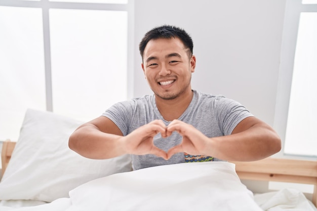 Young chinese man doing heart gesture sitting on bed at bedroom