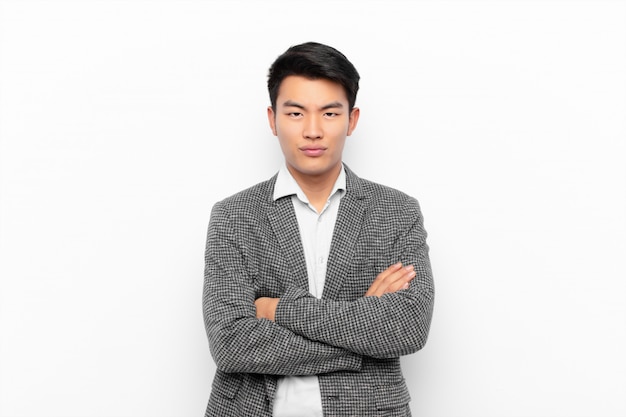 young chinese man feeling displeased and disappointed, looking serious, annoyed and angry with crossed arms against flat color wall