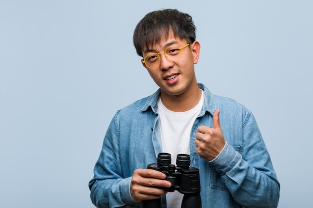 Young chinese man holding a binoculars smiling and raising thumb up