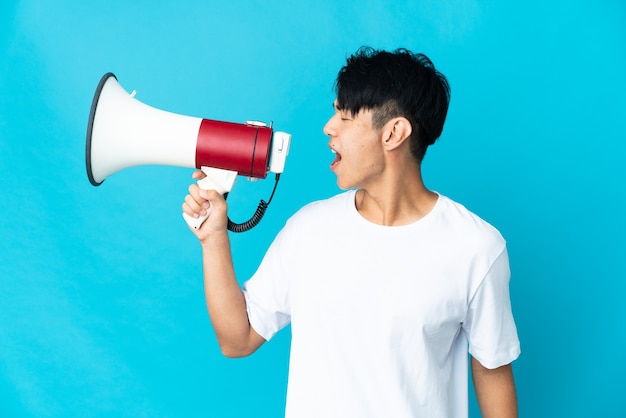 Young Chinese man isolated on blue wall shouting through a megaphone to announce something in lateral position