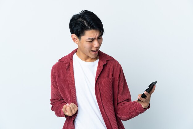 Young Chinese man isolated on white space using mobile phone and doing victory gesture