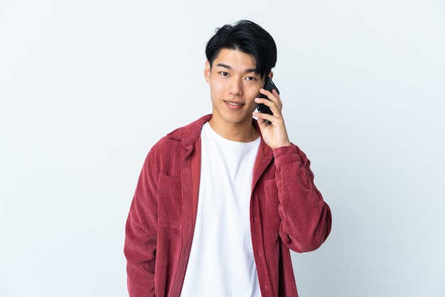 Young Chinese man isolated on white wall keeping a conversation with the mobile phone with someone