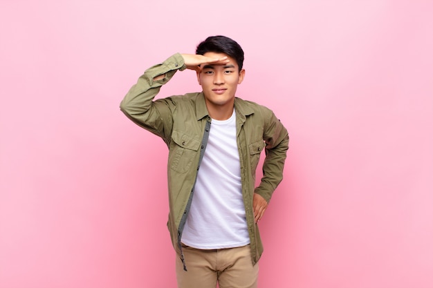 Young chinese man looking bewildered and astonished, with hand over forehead looking far away, watching or searching against flat color wall