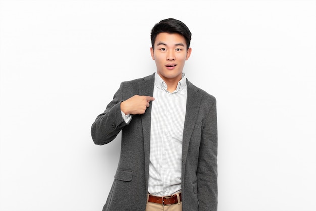 Young chinese man looking happy, proud and surprised, cheerfully pointing to self, feeling confident and lofty on flat color wall