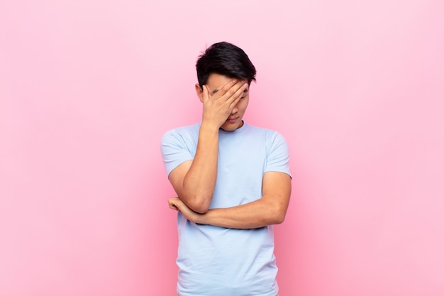 Young chinese man looking stressed, ashamed or upset, with a headache, coning face with hand on flat color wall