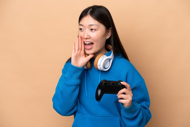 Young Chinese man playing with a video game controller isolated on beige background shouting with mouth wide open to the side