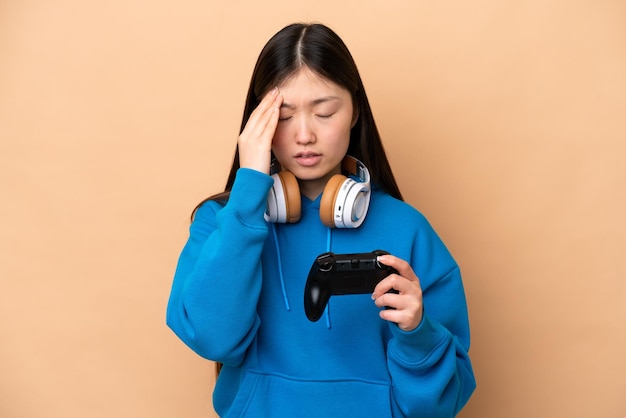 Young Chinese man playing with a video game controller isolated on beige background with headache