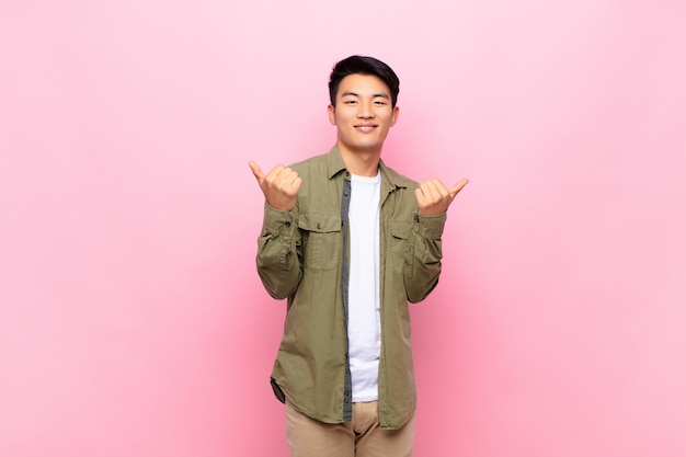 Young chinese man smiling joyfully and looking happy, feeling carefree and positive with both thumbs up against flat color wall