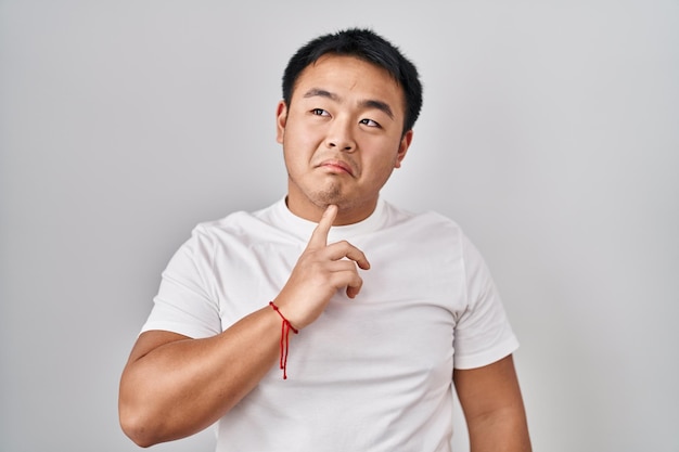 Young chinese man standing over white background thinking concentrated about doubt with finger on chin and looking up wondering