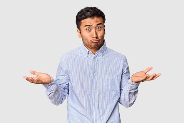 Young Chinese man in studio background doubting and shrugging shoulders in questioning gesture