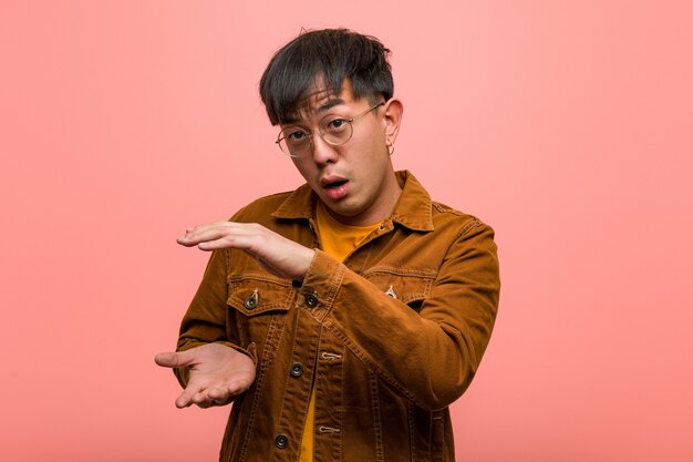 Young chinese man wearing a jacket holding something very surprised and shocked