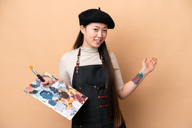 Young chinese painter woman isolated on beige background\
extending hands to the side for inviting to come