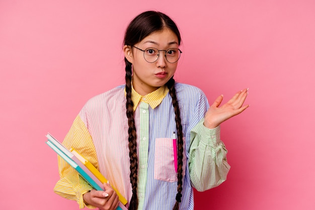 Photo young chinese student woman holding books wearing a fashion multicolour shirt and braid, isolated on pink background shrugs shoulders and open eyes confused.