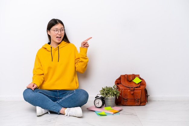 Молодая китайская студентка сидит на полу изолированно на белой стене, указывая пальцем в сторону и представляя продукт