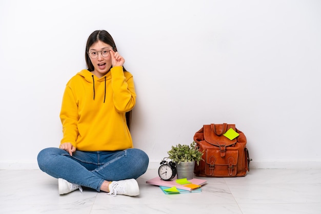 Young Chinese student woman sitting on the floor isolated on white wall with glasses and surprised