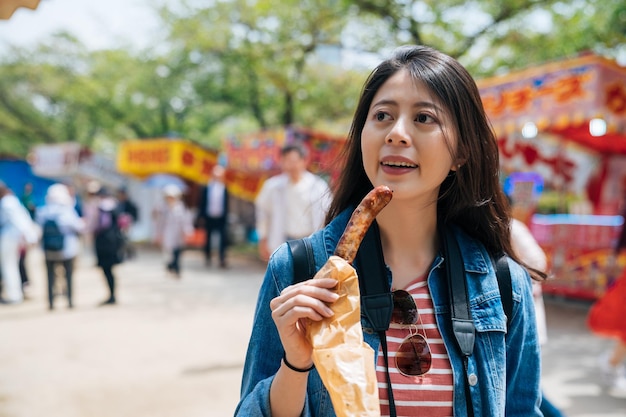 Il giovane viaggiatore cinese si unisce alla fiera del tempio di sakura nella menta giapponese. salsiccia d'acquisto del turista della ragazza dal venditore mobile della cucina. signora elegante che mangia cibo di strada nel festival di primavera giapponese.