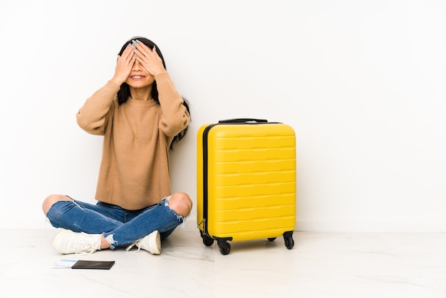 Young chinese traveler woman siting on the floor with a suitcase isolated covers eyes with hands, smiles broadly waiting for a surprise.