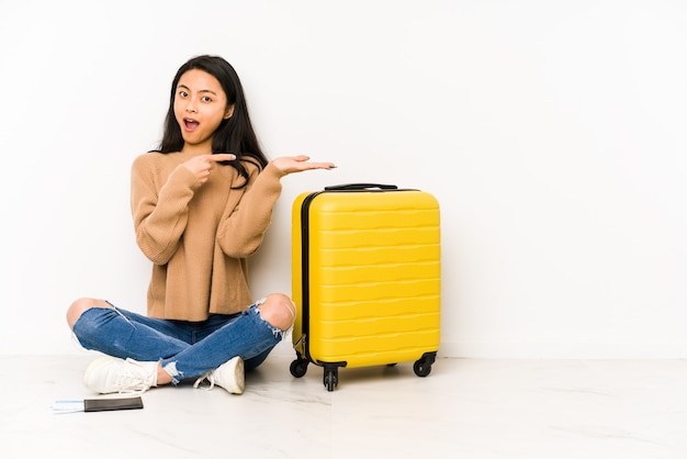 Young chinese traveler woman sittting on the floor with a suitcase excited holding a blank space on palm.