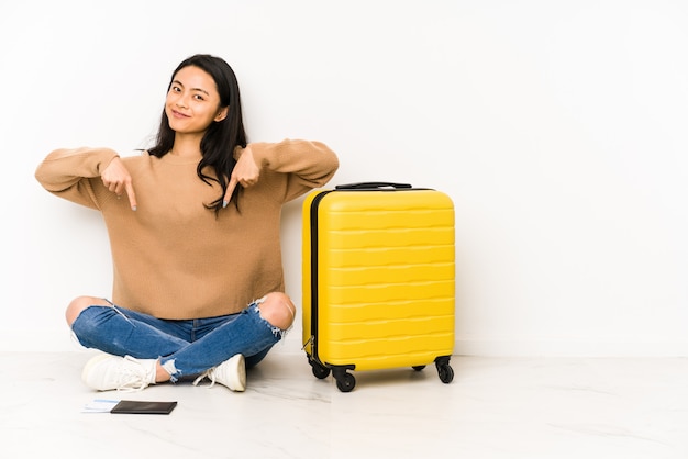 Young chinese traveler woman sittting on the floor with a suitcase points down with fingers