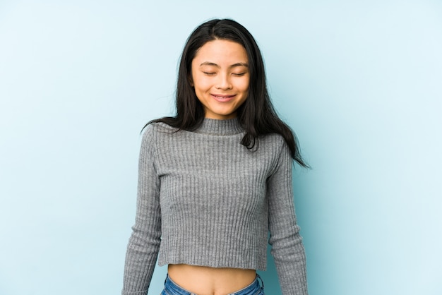 Young chinese woman on a blue wall laughs and closes eyes, feels relaxed and happy.