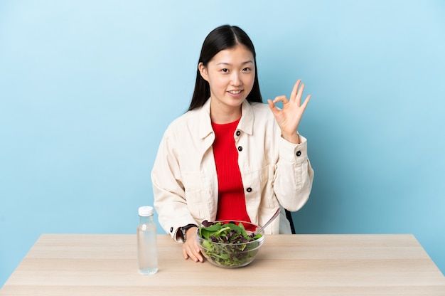 Giovane donna cinese che mangia un'insalata che mostra segno giusto con le dita