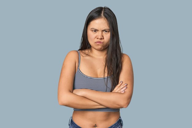 Young Chinese woman in gray top studio frowning face in displeasure keeps arms folded