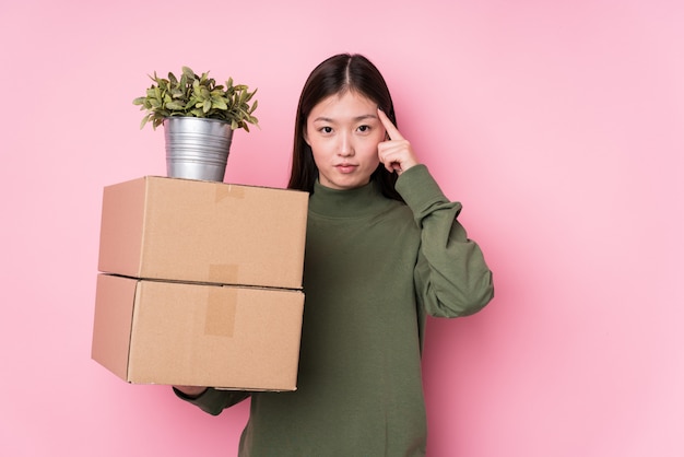 Молодая китайская женщина держа коробки изолировала указывать висок с пальцем, думая, сфокусированный на задаче.