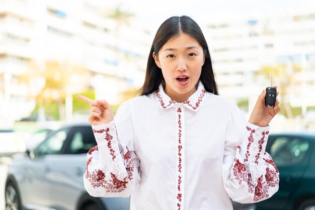 屋外で車のキーを保持している若い中国人女性が驚いて側を指している