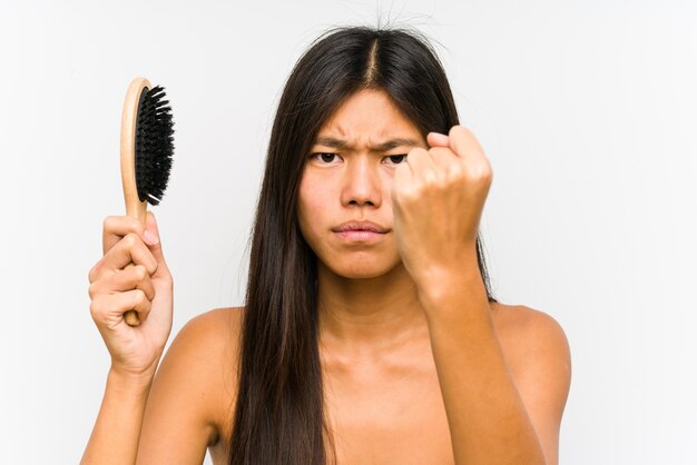Giovane donna cinese che tiene una spazzola per capelli isolata che mostra pugno, espressione facciale aggressiva.